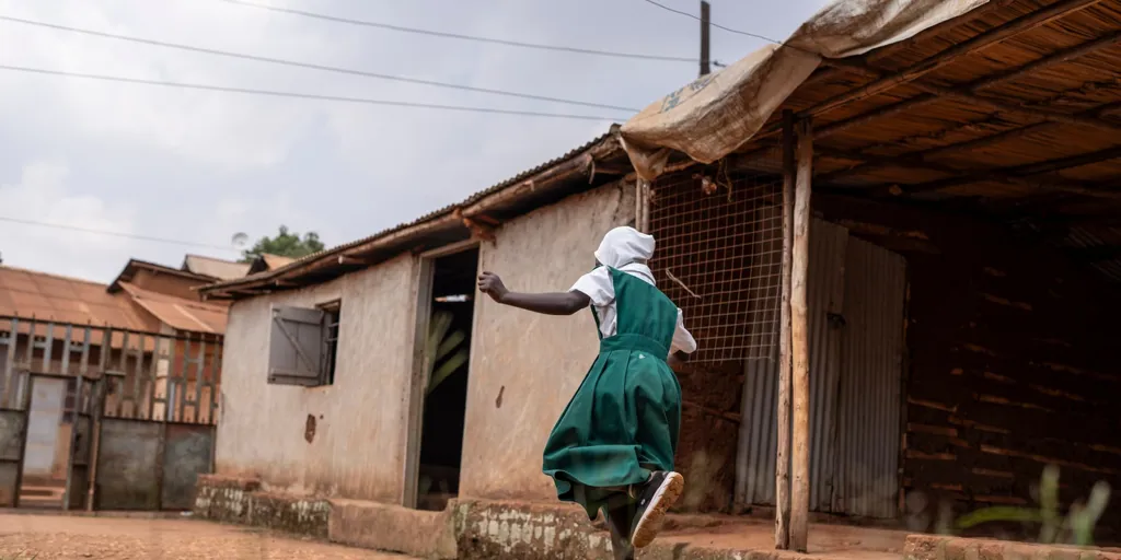 A refugee girl in a green school uniform plays a game in the yard of her home in Kampala, Uganda